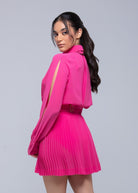Model wearing a bright pink Fierce mini dress with pleated skirt, slit long sleeves, and belted waist, three-quarter back view.