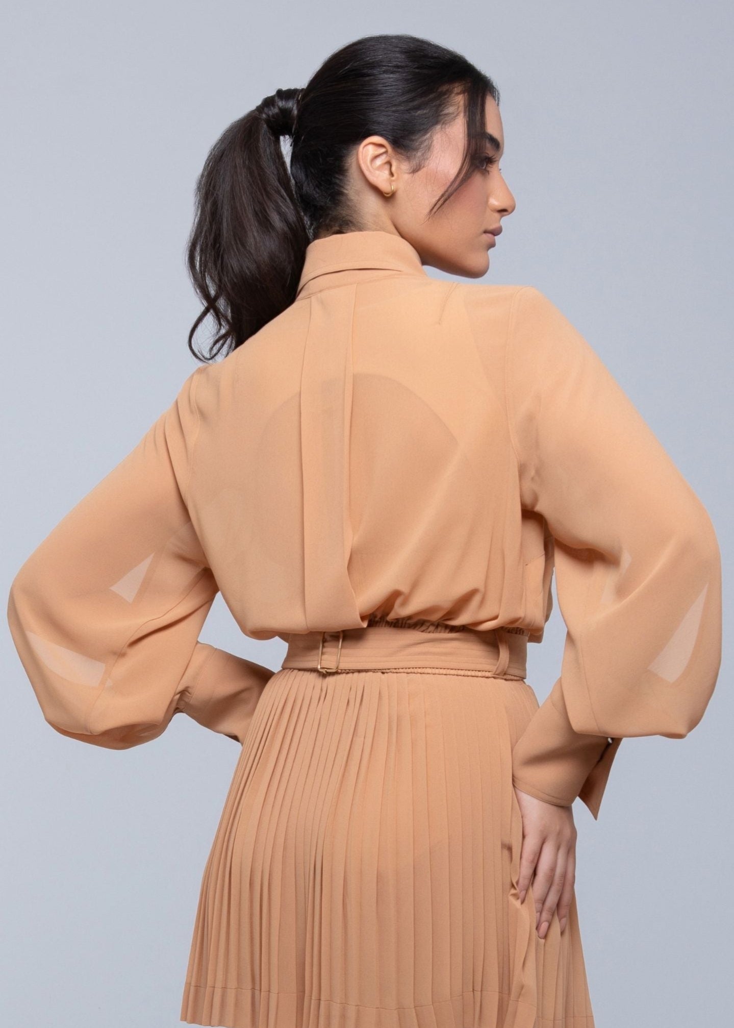 Woman wearing a camel-colored pleated Fierce skirt and sheer long-sleeve blouse, posing with her back to the camera and head turned to the side.