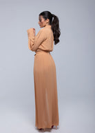 Woman wearing a camel-colored pleated Fierce maxi dress with long sleeves, posing from the side with hands near her face.