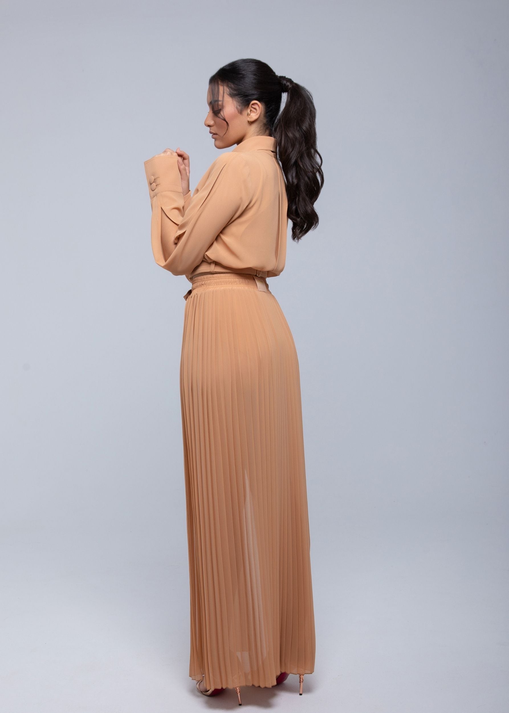 Woman wearing a camel-colored pleated Fierce maxi dress with long sleeves, posing from the side with hands near her face.
