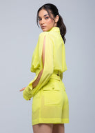 Model in a neon yellow Fierce long-sleeve blouse with open slit sleeves, paired with matching high-waist tailored shorts, looking over her shoulder.