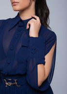 Close-up of a model in a Fierce sheer navy blue blouse featuring fabric-covered buttons, a pointed collar, a decorative chest pocket, and a wide cuff detail with button accents, paired with a gold-buckled belt.