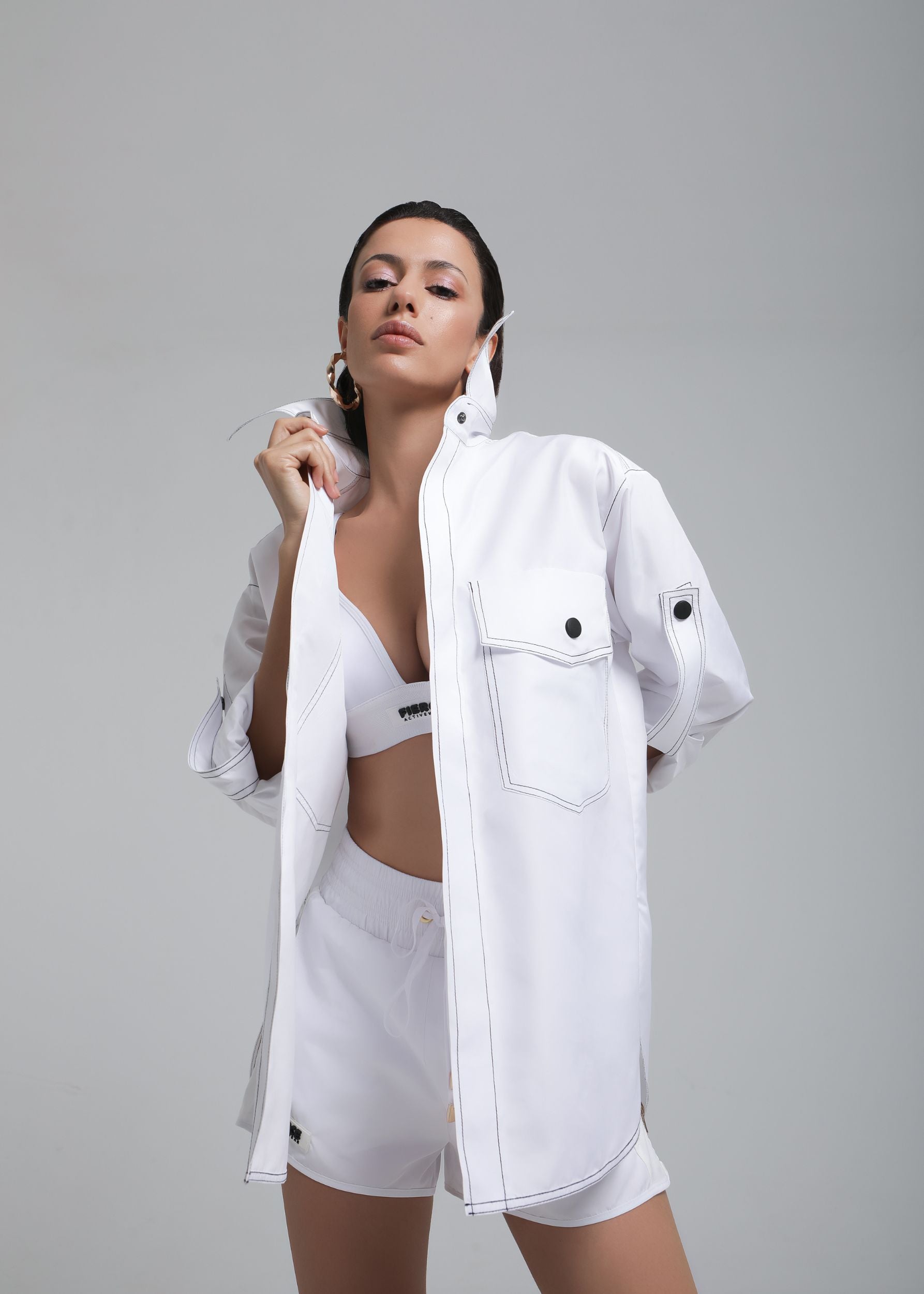 Confident model wearing Fierce all-white set featuring Aero Chemise with contrast stitching, sports bra, and shorts – front pose with one hand on collar