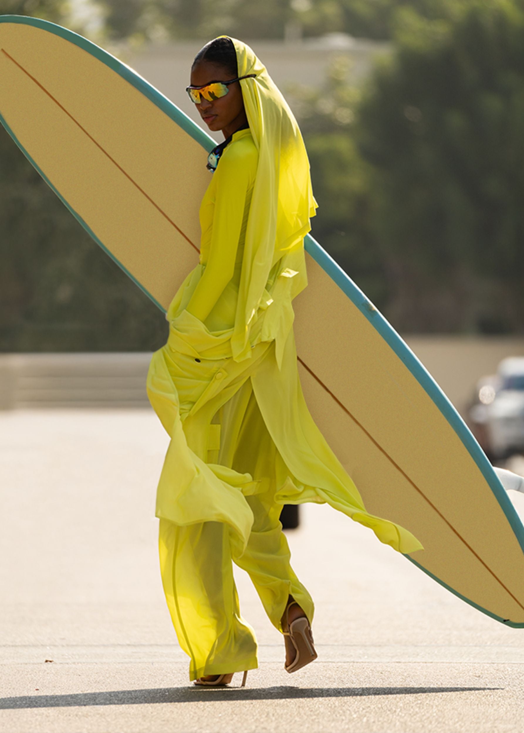 Person in a bright Fierce yellow outfit holding a surfboard on a road.